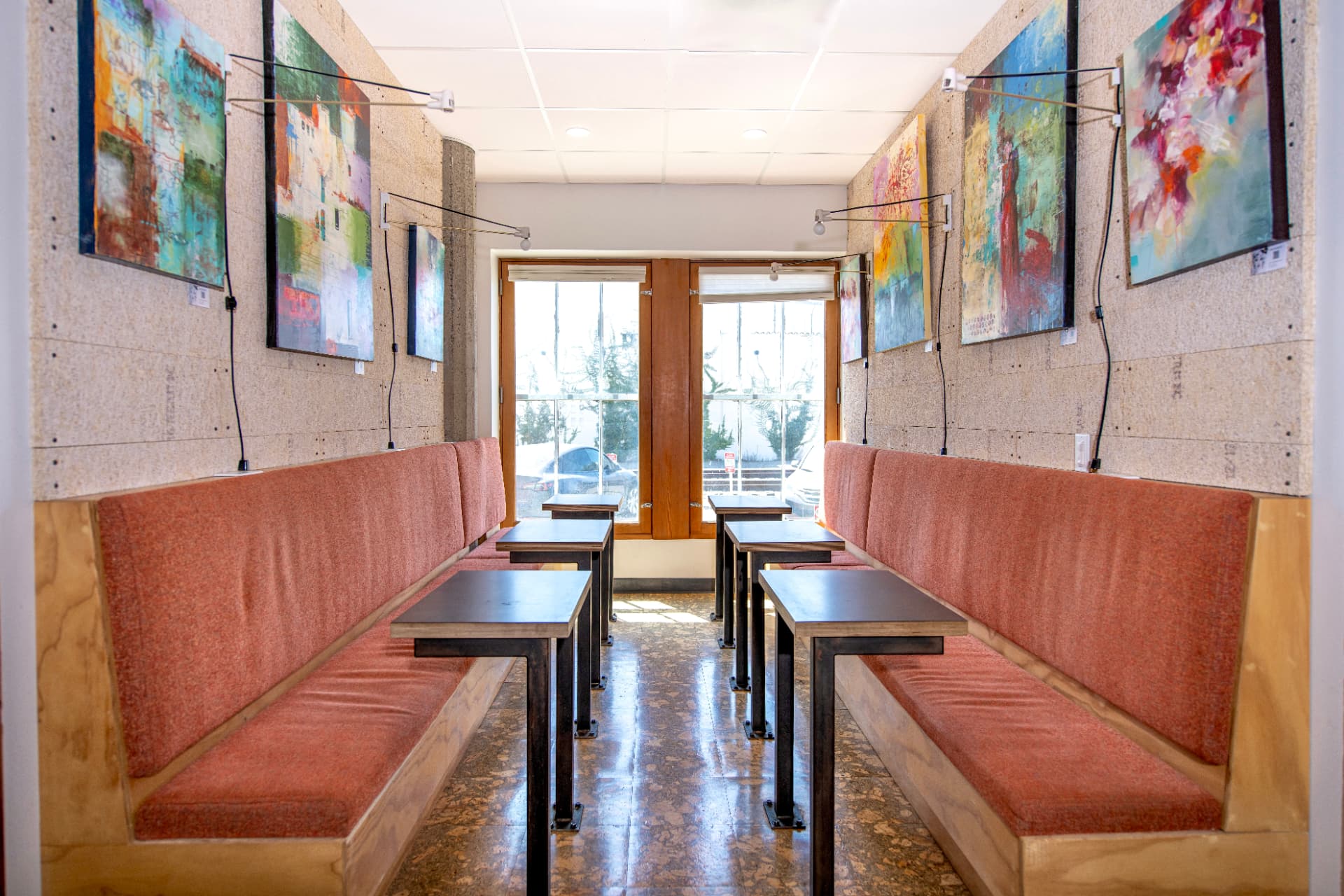 Coworking booth alcove at Kiva Funk Zone with built-in wood banquettes, rust-orange cushions, workstation tables and colorful abstract paintings