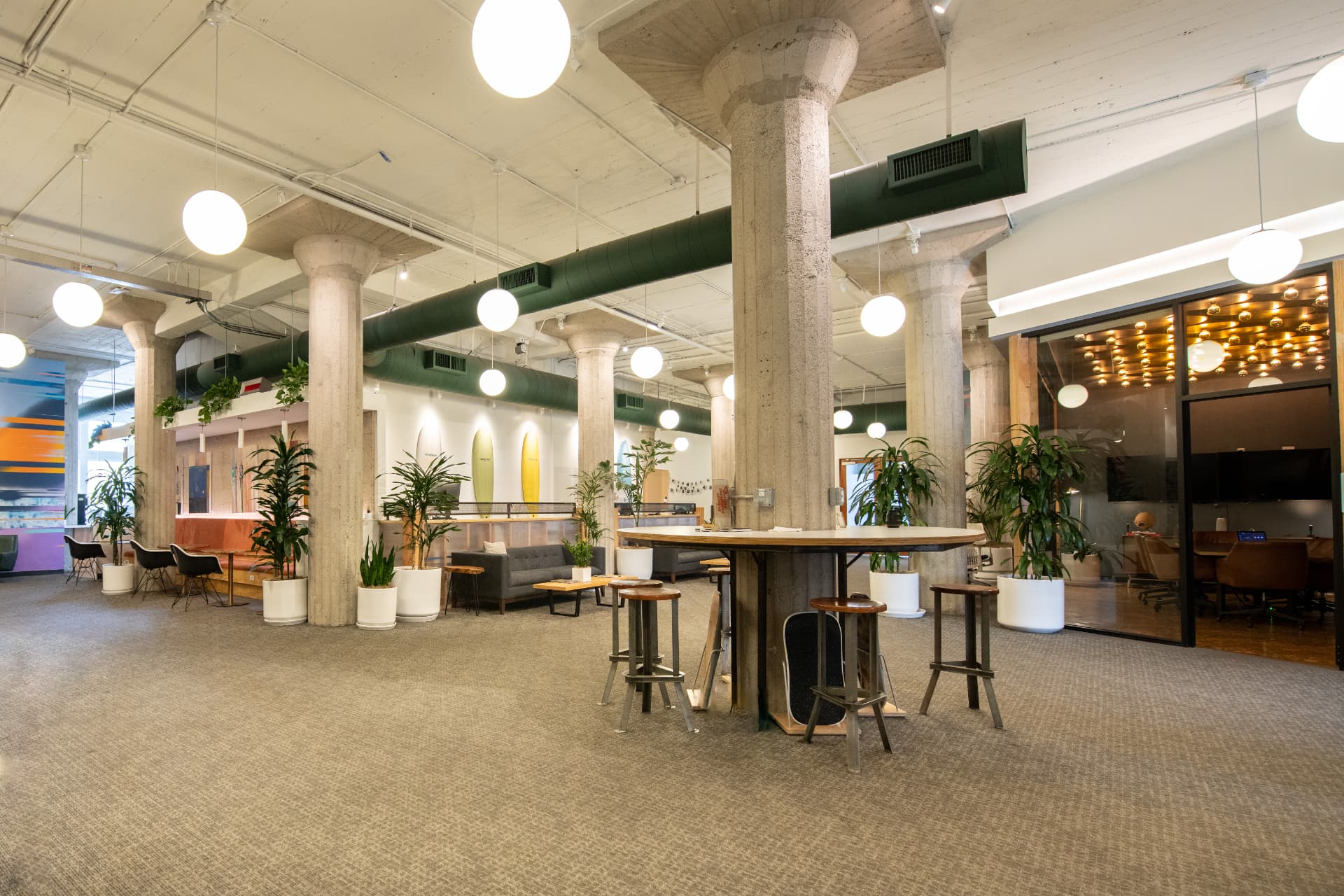 Open coworking lounge at Kiva Funk Zone with concrete pillars, a round high-top table, lounge sofa, wall-mounted surfboards and a glass conference room